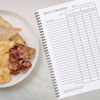 Daily Macro Nutrients Tracker journal next to a plate of breakfast toast scrambled eggs and bacon